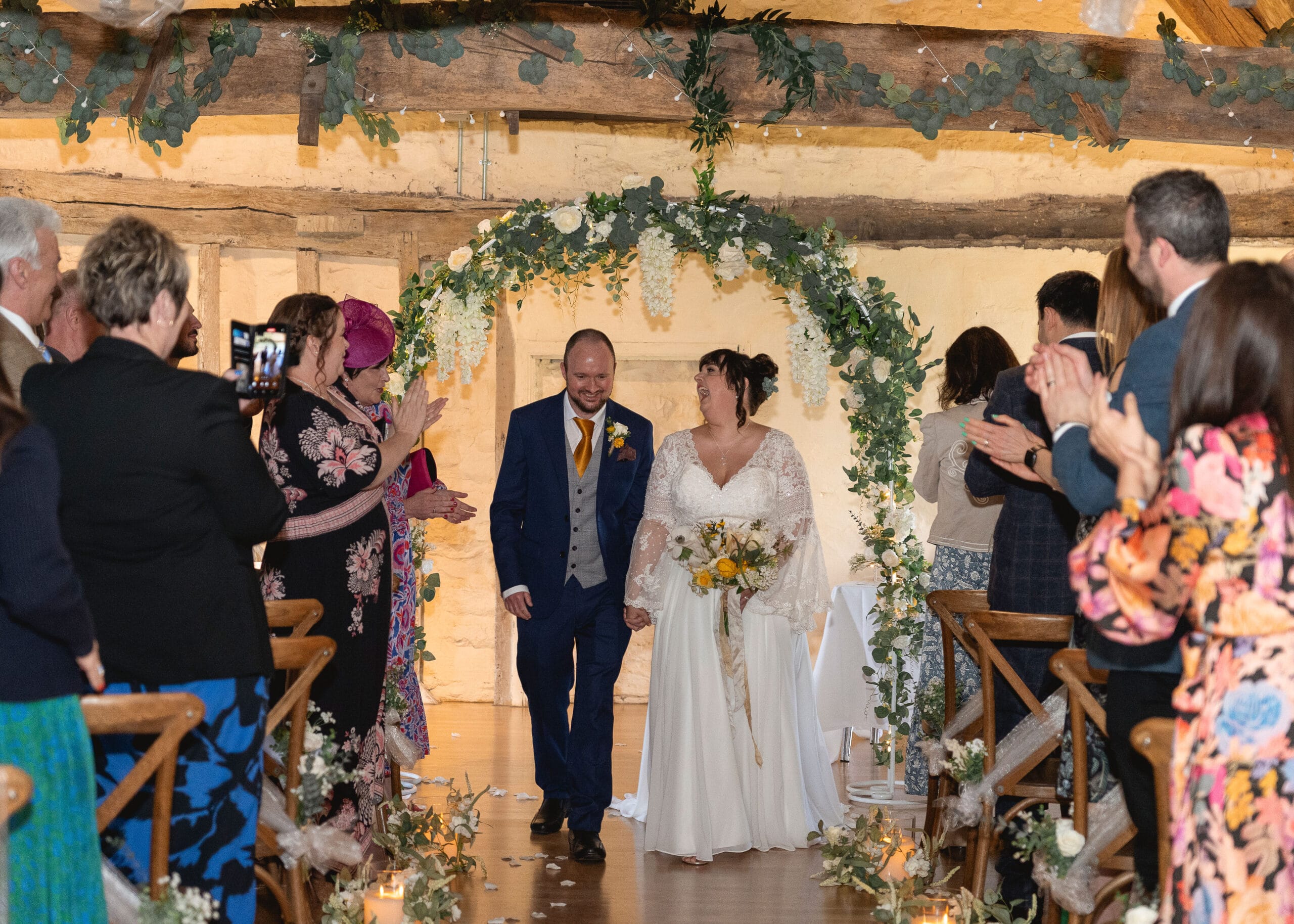 Bride (right) and Groom (Left) walk down an aisle after getting married in the Lady Margaret Hall with a floral arch behind them